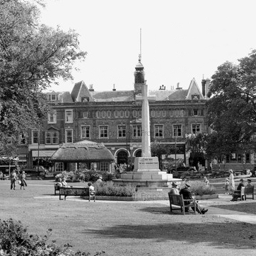 A black and white photo of a public square featuring a tall monument in the center, surrounded by benches and trees. In the background, there are historic buildings with ornate architecture.
