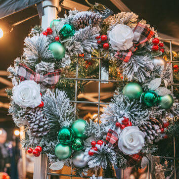 A festive holiday wreath featuring white roses, green and silver ornaments, pinecones, and red berries, designed with a frosted appearance.